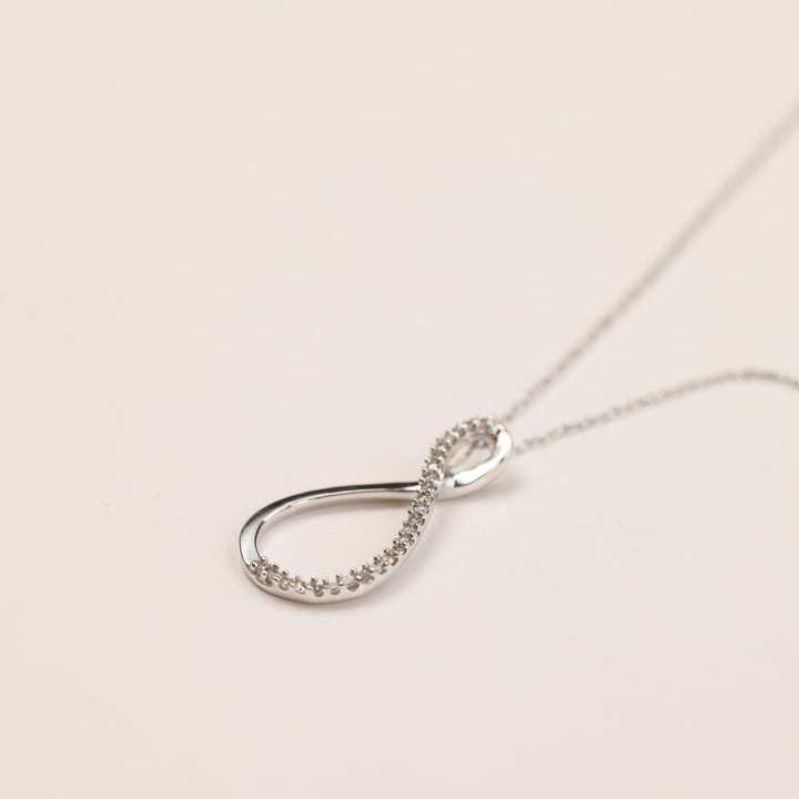 A silver infinity pendant with small embedded stones on a thin chain necklace, reminiscent of intricate jewelry like a 14 Karat Yellow Gold Tanzanite Diamond Ring - Size 5, against a plain background.