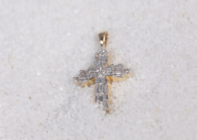 A 14 Karat Yellow Gold Diamond Cross Pendant resting on a bed of sand.