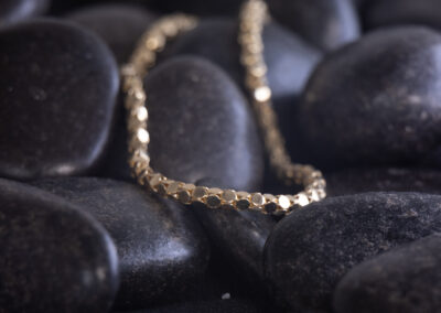 A gold chain is laying on top of black rocks, showcasing a 14 Karat Yellow Gold Cylindric Bracelet.