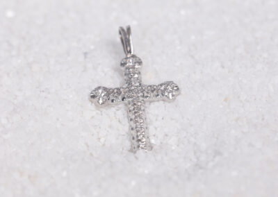 A silver cross pendant laying on top of sand, accompanied by a delicate 14 Karat Yellow Gold White Stone Tennis Bracelet.
