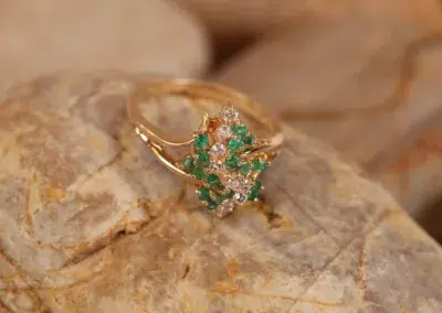 Gold ring with green gemstones and diamonds displayed on a rock, accompanied by a 14 Karat Yellow Gold Figaro 25" Chain.