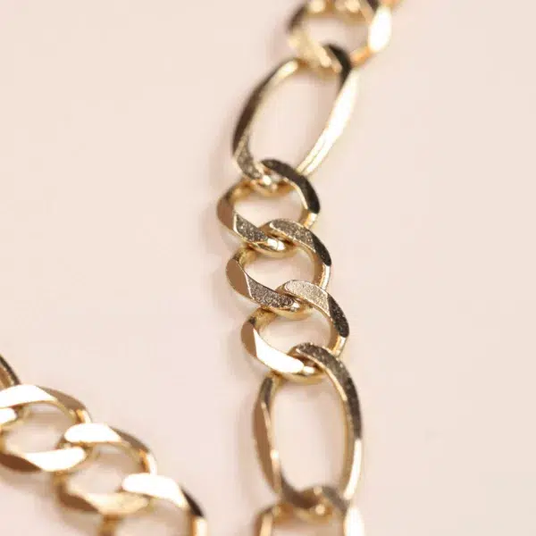 Close-up of a 14 Karat Yellow Gold Figaro 25" chain with interlocking links on a pastel background.