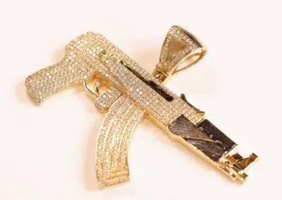 A golden pendant shaped like a gun, embellished with sparkling crystals, hangs from a 14 Karat Yellow Gold Figaro 25" Chain.