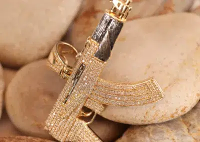 14 Karat Yellow Gold and Diamond-Encrusted Pendant in the Shape of a Gun resting on smooth stones, attached to a 14 Karat Yellow Gold Figaro 25" Chain.