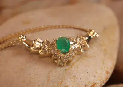 Gold bracelet with an emerald centerpiece and diamond accents displayed on a stone, featuring a 14 Karat Yellow Gold Figaro 25" Chain.