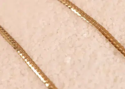 Close-up of two 14 Karat Yellow Gold Figaro 25" chains on a textured beige surface.
