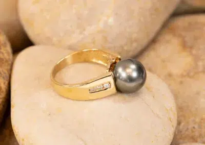 Gold ring with a large pearl and a 14K YG Tanzanite & Diamond Pendant, resting on a smooth stone among other pebbles.