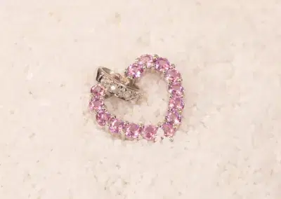 A heart-shaped ring with pink gemstones and a 14K YG Tanzanite & Diamond Pendant, resting on a sandy surface.