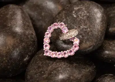 A heart-shaped pink 14K YG Tanzanite & Diamond pendant rests atop smooth, dark stones.