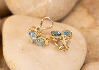 A pair of 10 Karat Yellow Gold Ram Ring earrings with blue gemstones resting on a smooth, beige stone.