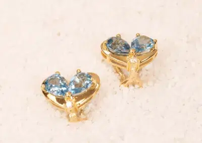 A pair of 10 Karat Yellow Gold earrings with heart-shaped blue gemstones set on a white sandy texture.