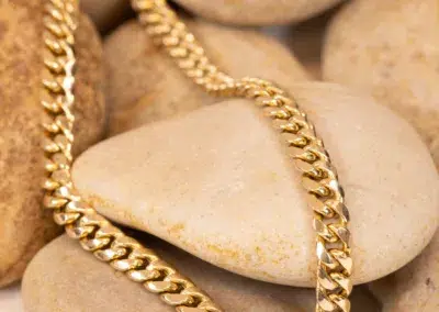 A gold chain necklace draped over smooth, beige stones showcases the 10 Karat Yellow Gold Ram Ring.