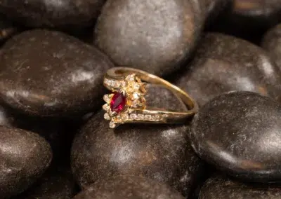 10 Karat Yellow Gold Ram ring with a central red gem and small diamonds embedded around it, resting on a bed of smooth, dark pebbles.
