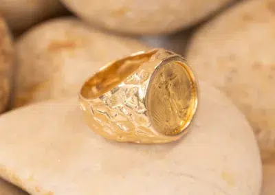 A 10 Karat Yellow Gold Ram Ring with an embossed design rests on smooth, pale stones.