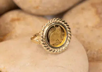 10 Karat Yellow Gold Ram Ring with an embedded coin, featuring a male profile, displayed on a smooth, light-colored stone.