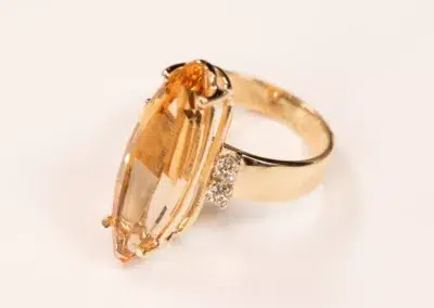 14K YG Diamonds & Peach Tourmaline Ring featuring a large, oval-shaped peach tourmaline gemstone flanked by small diamonds, set against a white background.