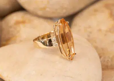 14K YG Diamonds & Peach Tourmaline Ring with a large, vertical peach tourmaline gemstone and small diamonds, displayed on smooth pebbles.