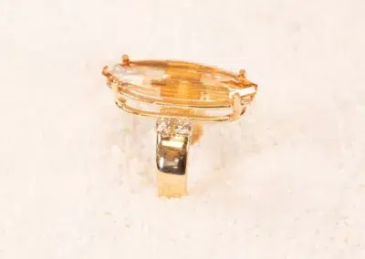A large, oval-shaped amber gemstone set in a 14K YG Diamonds & Peach Tourmaline ring, displayed on a white grainy surface.