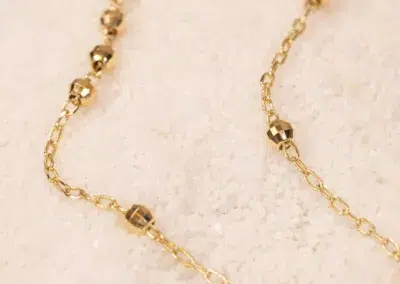 Close-up image of a delicate gold necklace with small spherical beads and a 14K YG Diamonds & Peach Tourmaline Ring, resting on a textured pale pink surface.