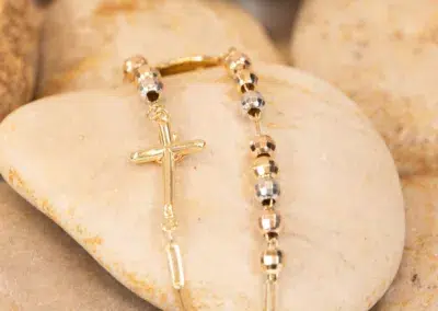 Gold crucifix pendant on a beaded bracelet, resting on smooth, multi-toned pebbles adorned with 14K YG Diamonds & Peach Tourmaline Ring.