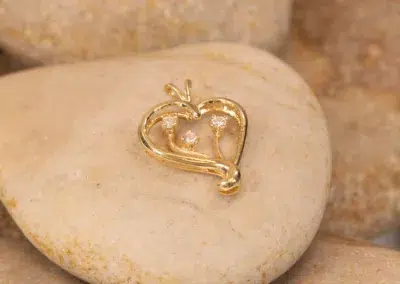 A gold heart-shaped pendant with three embedded diamonds, resting on smooth beige 14K YG Diamonds & Peach Tourmaline Ring.