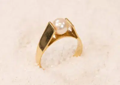 A 14K YG Diamonds & Peach Tourmaline ring with a large pearl set on top, placed on a sandy surface.